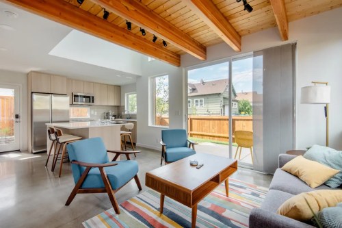 Bright, modern living room with wooden beams, a cozy sofa, chairs, and a view of the outdoor patio through large windows.