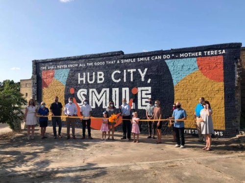 A group of people stands in front of a colorful mural reading "Hub City Smile," holding a ribbon for a ceremony.