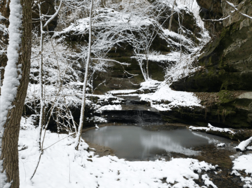 A serene winter scene with a snowy landscape, a small pond, and rocky cliffs surrounded by bare trees.