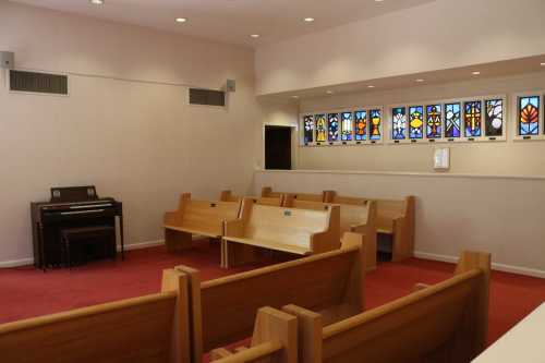 Interior of a small chapel with wooden benches, a keyboard, and colorful stained glass windows.