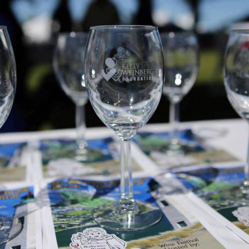 Clear wine glasses with the Kelly Weinberg Foundation logo, placed on a table with festival brochures.