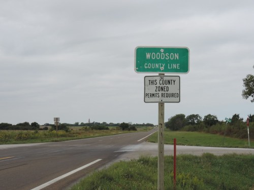 Sign marking the Woodson County line, with an additional sign about zoning and permit requirements. Overcast sky in background.