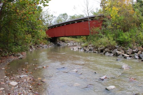 A red covered bridge spans a shallow creek surrounded by trees and rocky banks in a serene natural setting.