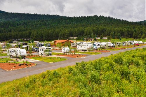 A scenic RV park with several parked trailers surrounded by green trees and mountains under a cloudy sky.