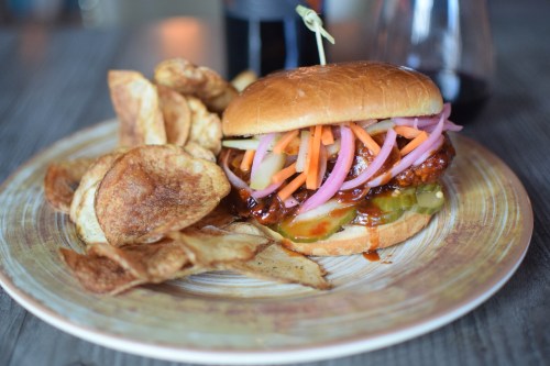 A delicious burger topped with pickles, carrots, and onions, served with crispy potato chips on a wooden plate.