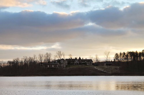 A serene lake scene with a large house on a hill, surrounded by trees under a cloudy sky at sunset.