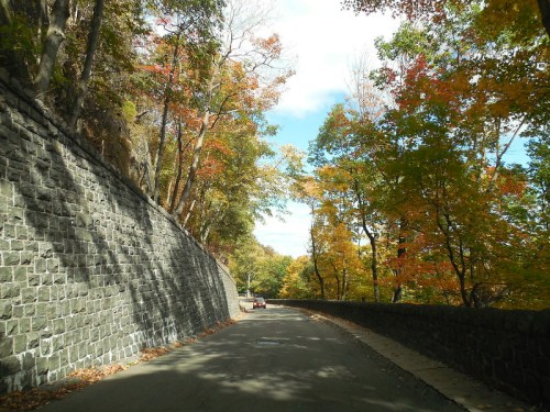 A scenic road lined with colorful autumn trees and a stone wall under a partly cloudy sky.