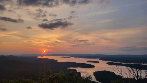 A serene sunset over a lake, with islands and lush green hills under a colorful sky filled with clouds.