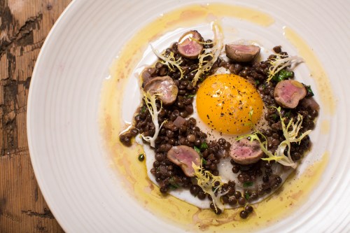 A plate of lentils topped with a poached egg, sliced meat, and garnished with greens, drizzled with olive oil.