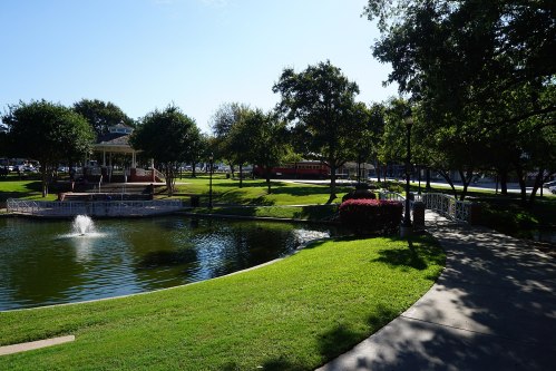 A serene park scene featuring a pond, fountain, green lawns, trees, and a walkway under a clear blue sky.