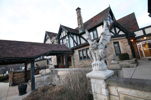 A stone building with a steep roof and decorative gargoyle statue in front, surrounded by landscaping.