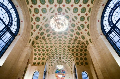 Ornate ceiling with colorful patterns and a chandelier, viewed from below in a grand architectural space.