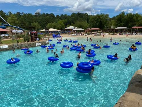 A busy water park scene with people floating on blue inner tubes in a clear pool under a sunny sky.