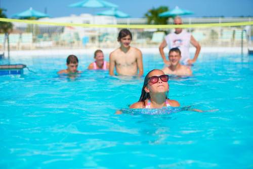 A girl in sunglasses floats in a pool, while several others watch and enjoy the sunny day.