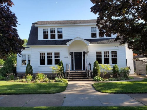 A charming two-story white house with a black door, surrounded by greenery and a well-maintained lawn.