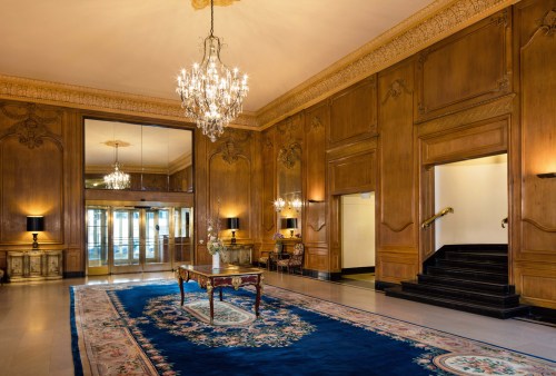 Elegant lobby with wooden walls, chandeliers, and a blue rug, featuring a table and mirrored entrance.