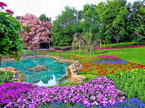A vibrant garden with colorful flowers, a clear pond, and lush greenery, set against a rocky backdrop.