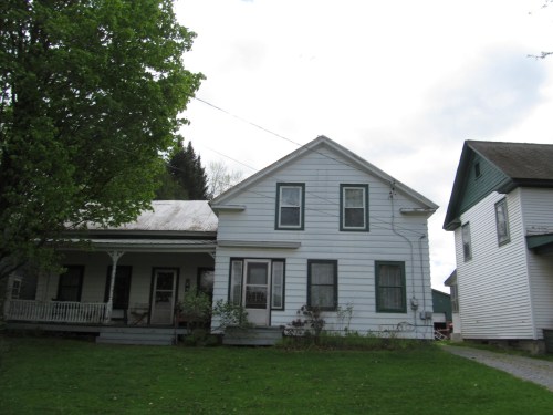A white two-story house with green trim, surrounded by trees and grass, featuring a porch and a neighboring building.