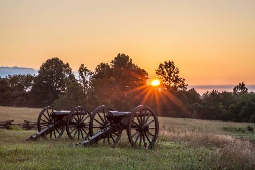 Two historic cannons sit in a grassy field at sunrise, with trees silhouetted against the colorful sky.