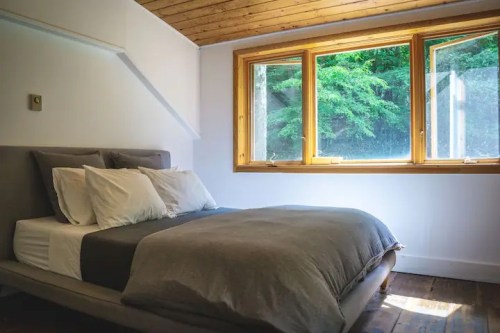 Cozy bedroom with a gray bedspread, white pillows, and large windows overlooking greenery.