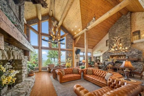 Spacious rustic living room with leather sofas, stone fireplace, large windows, and wooden beams, surrounded by nature.