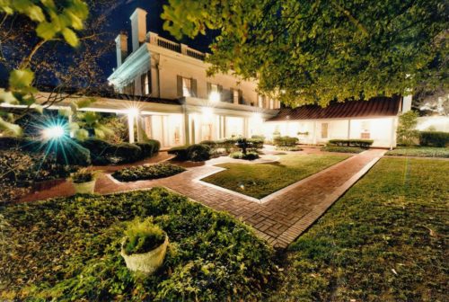 A beautifully lit historic house at night, surrounded by a well-maintained garden and pathways.