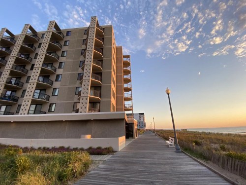 A boardwalk lined with grass and a tall building at sunset, with a clear sky and scattered clouds.