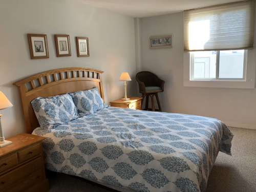 Cozy bedroom with a wooden bed, blue patterned bedding, nightstands, and a chair by the window. Soft lighting.