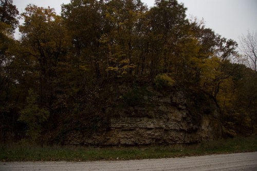 A rocky outcrop covered with trees and autumn foliage, set against a cloudy sky and gravel road.