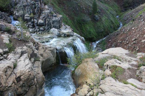 A serene waterfall cascades over rocky terrain, surrounded by lush green hills and a winding stream.