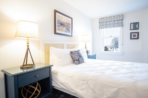 Cozy bedroom with a white bed, blue nightstands, lamps, and artwork, featuring natural light from a window.