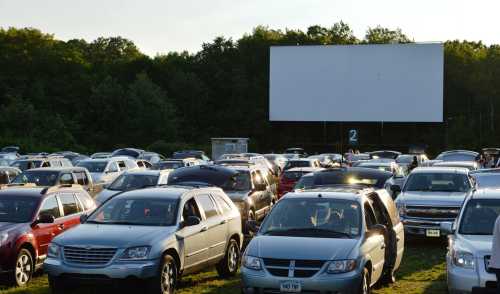 A crowded drive-in theater parking lot with cars facing a large outdoor movie screen. Trees in the background.