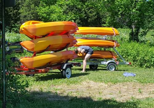A person adjusts a kayak on a trailer, with stacked orange kayaks in the background and greenery surrounding the area.