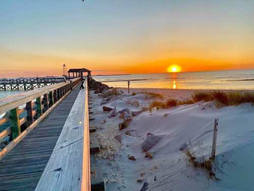 A wooden pier extends towards a vibrant sunset over calm waters, with sandy dunes and gentle waves in the foreground.