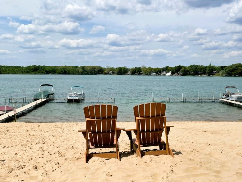 Two wooden chairs on a sandy beach facing a calm lake with boats and trees in the background under a cloudy sky.
