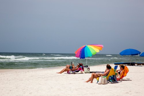 People relaxing on a sandy beach under colorful umbrellas, with gentle waves in the background.