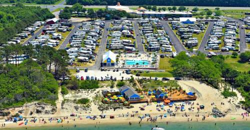 Aerial view of a beachside RV park with a pool, surrounded by campers and beachgoers enjoying the sandy shore.