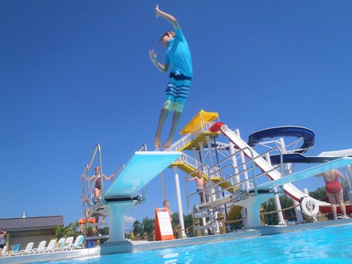 A child jumps off a diving board into a pool, with water slides and people in the background on a sunny day.