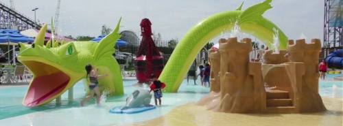 Children play in a water park featuring a large green dragon slide and a sandcastle structure.