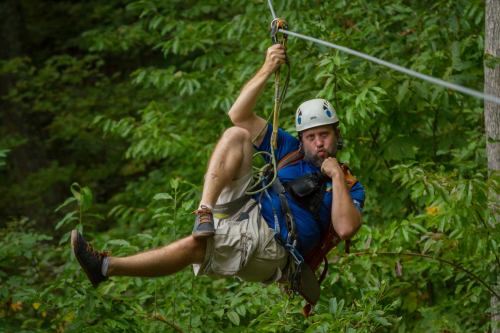 A person in a helmet zip-lining through a forest, striking a playful pose while hanging from the cable.