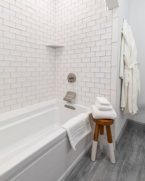 A clean bathroom with a white tiled bathtub, stacked towels, and a wooden stool beside a plush bathrobe hanging on the wall.