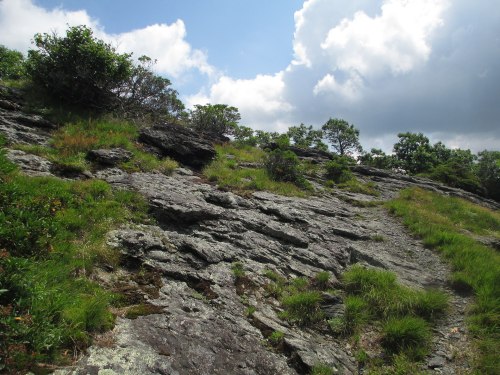 Rocky hillside with patches of grass and shrubs under a partly cloudy sky.