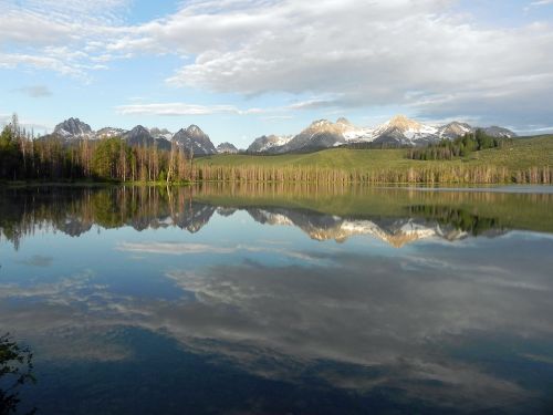 A serene lake reflects snow-capped mountains and a blue sky, surrounded by lush greenery and trees.
