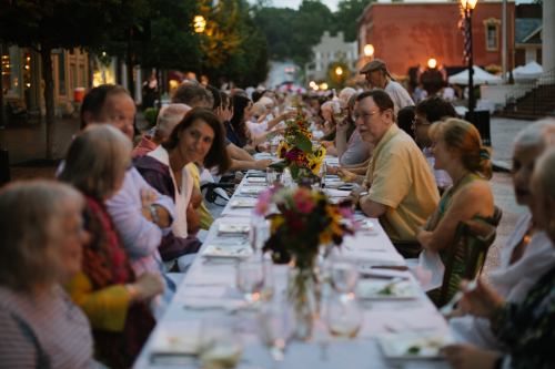 A long outdoor dining table filled with people enjoying a meal, surrounded by flowers and a festive atmosphere.