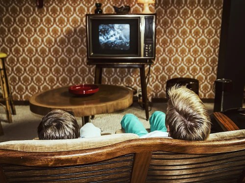 Two children sit on a vintage couch, watching an old television in a retro living room with patterned wallpaper.