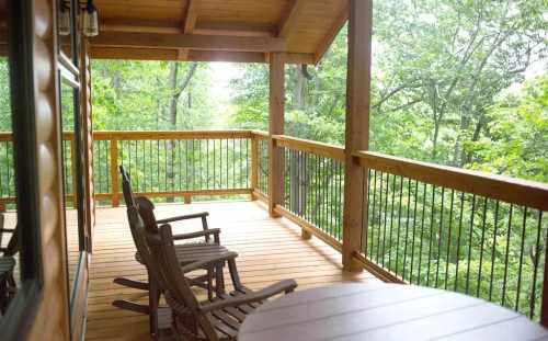 A wooden porch with rocking chairs overlooks a lush green forest. Natural light filters through the trees.