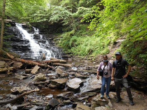 A couple stands by a waterfall in a lush green forest, surrounded by rocks and trees.