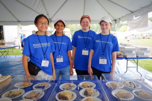 Four smiling volunteers in blue shirts stand behind a table with plates of food under a tent.