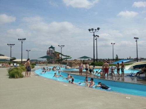 A busy water park with people enjoying a lazy river, water slides, and sunbathing areas under a cloudy sky.