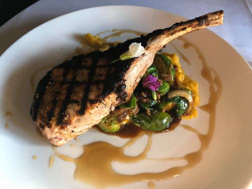 A grilled pork chop on a plate, served with Brussels sprouts and a drizzle of sauce, garnished with a flower.
